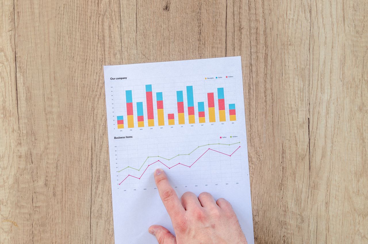 Services-01 A hand points to colorful business charts and graphs on a paper sheet on a wooden desk.